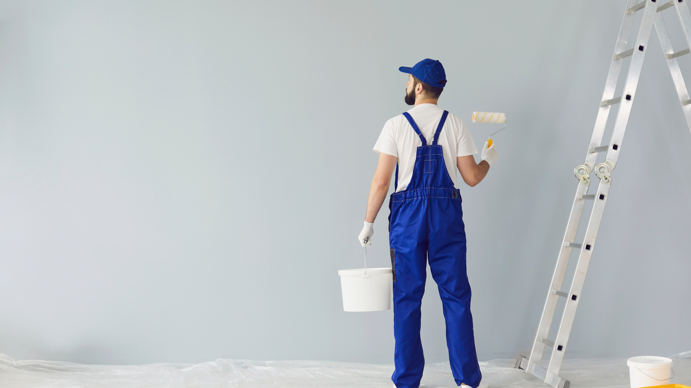 Painter in blue overalls holding a paint bucket and roller beside a ladder